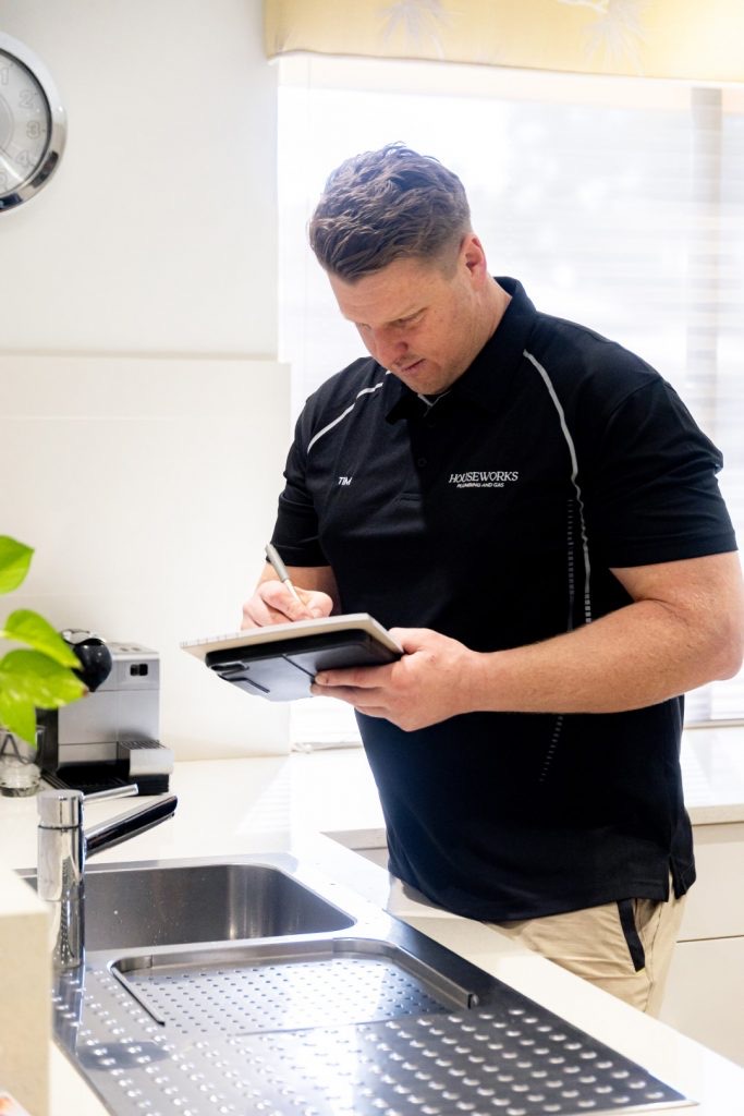 Plumber in a HouseWorks polo writing notes on a pad beside a stainless double sink